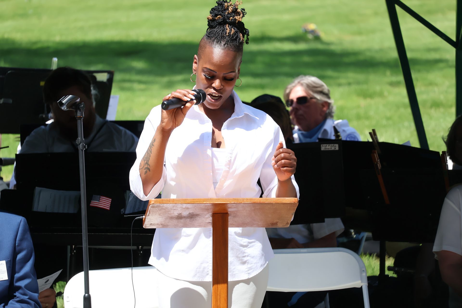 Woman in white speaking at a podium outdoors, with seated musicians in the background.