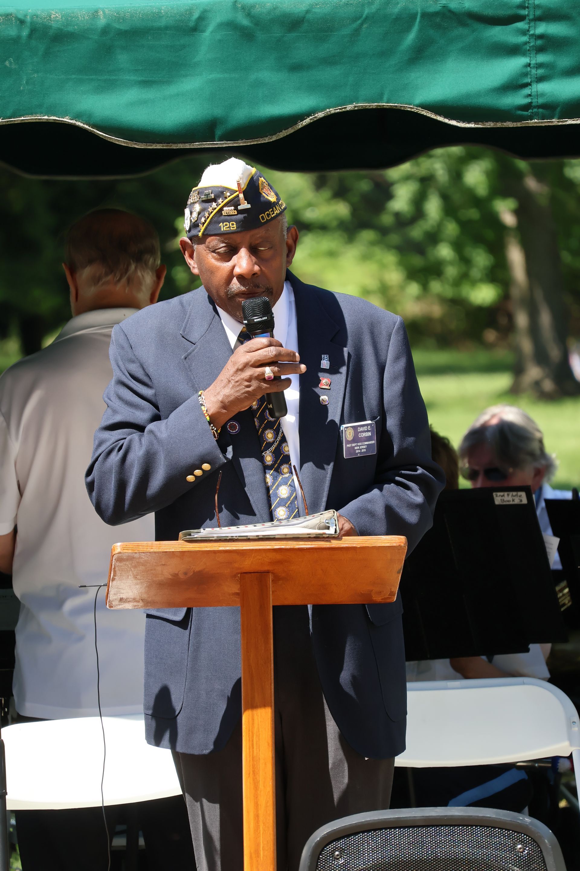 Man speaking at a podium under a green canopy outdoors, wearing a dark suit and patterned cap.