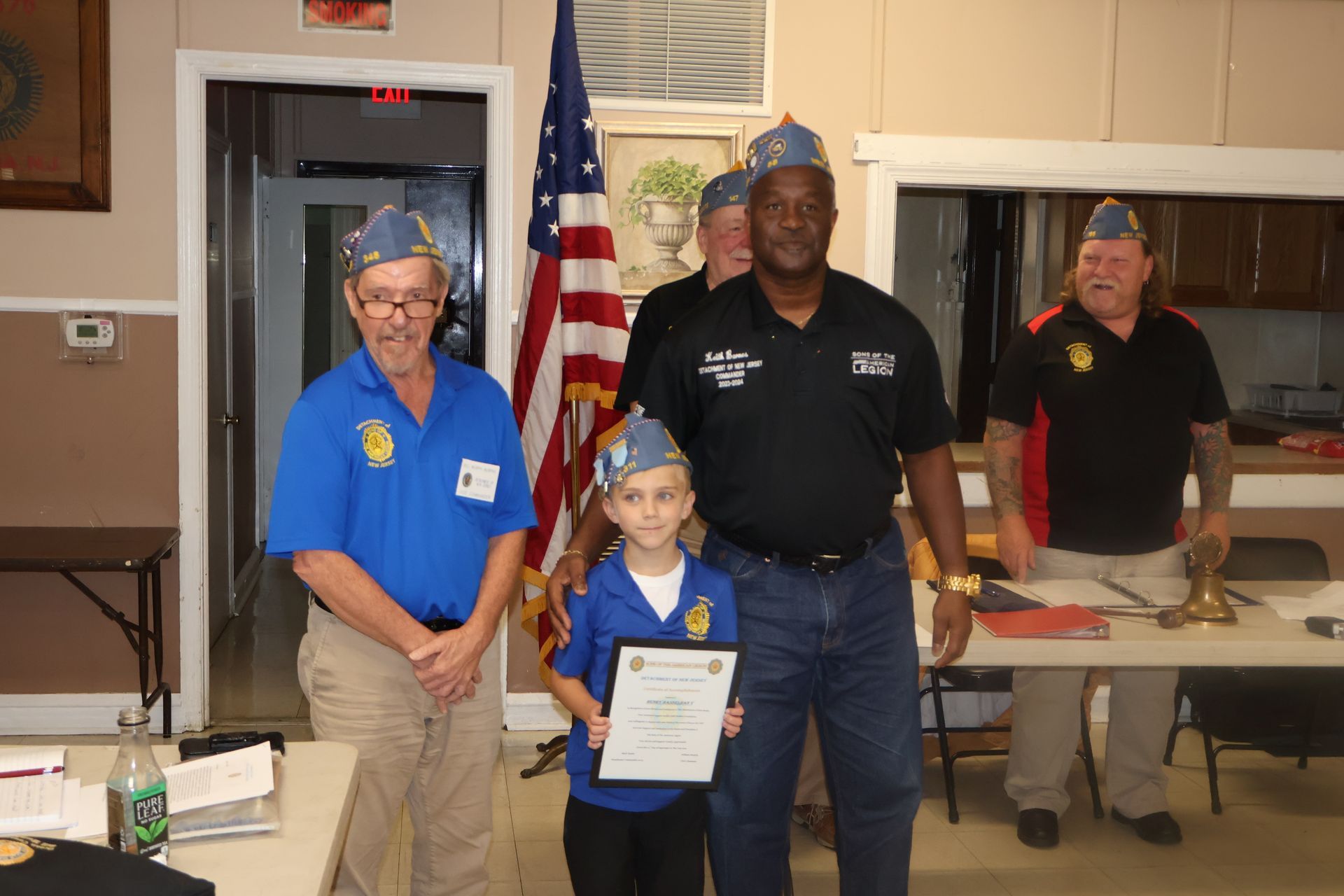 People in blue and black shirts stand near a child holding a certificate, next to an American flag.