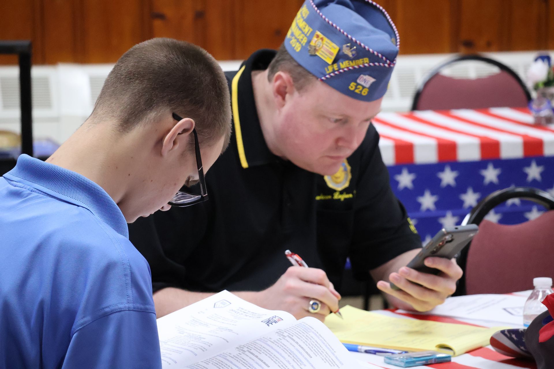 Two men at a table, one in a veteran's cap using a phone and pen, the other reviewing papers.