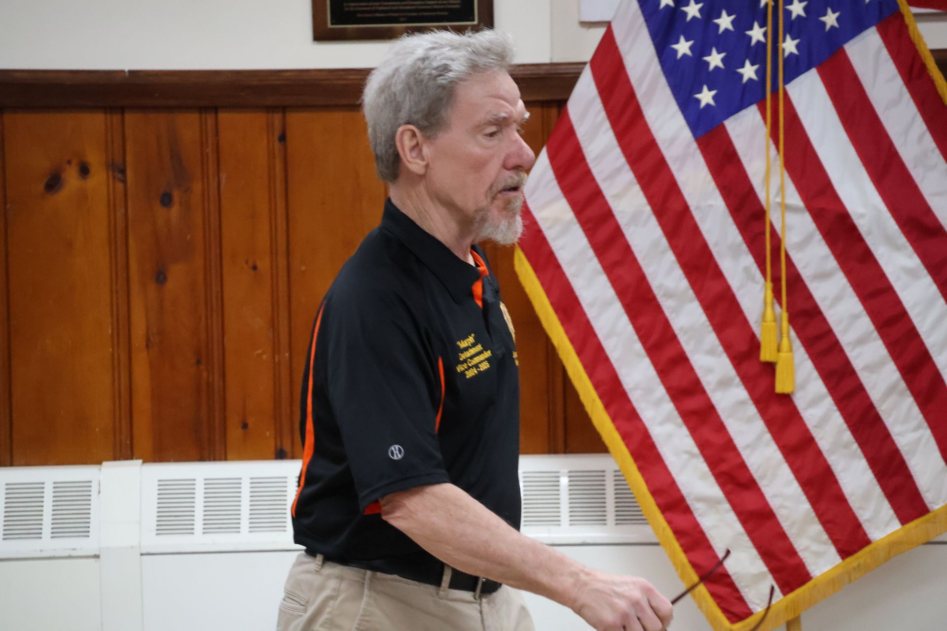 Man in black and orange shirt speaking in a room with American flag in background.