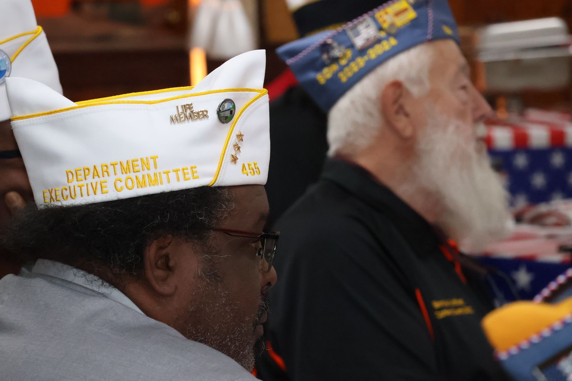 Two men wearing military-style hats at an event. The hats have text and emblems. One man has a long white beard.