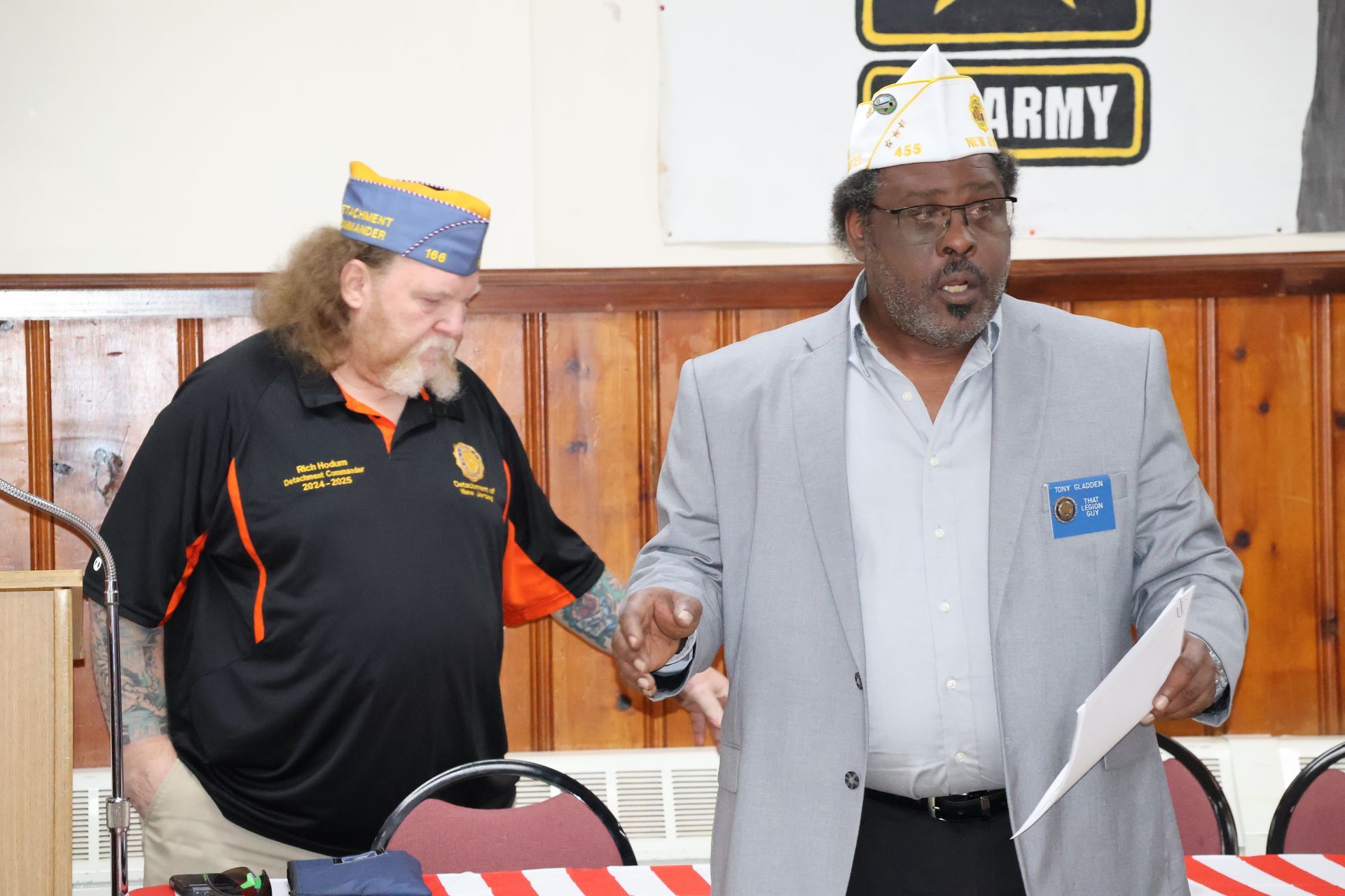 Man in gray suit speaks, holding paper; another man in black shirt stands nearby, both wearing decorated hats.