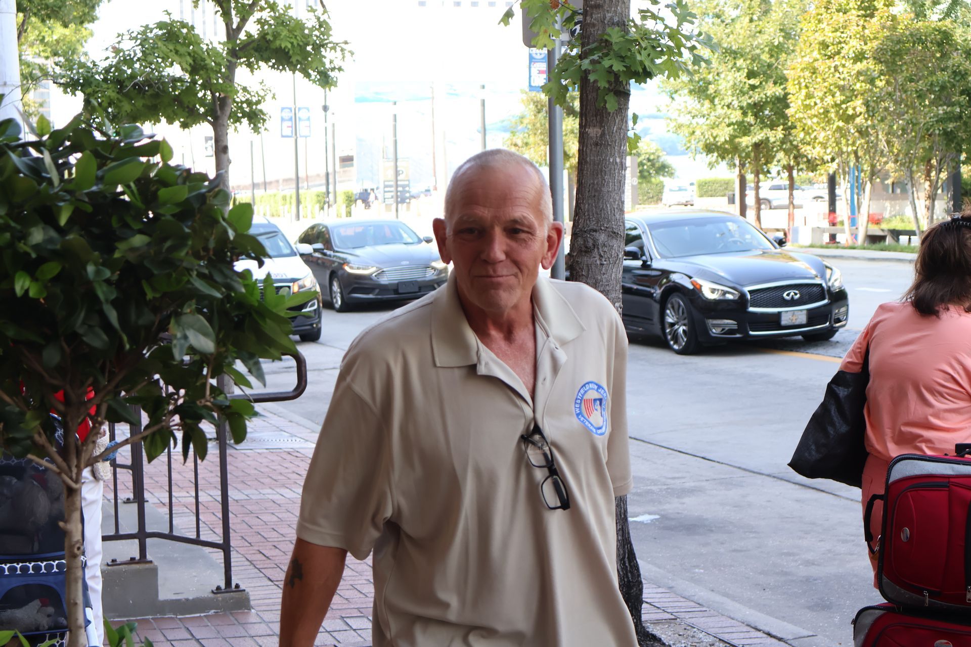Man in tan shirt with glasses smiles on sidewalk with cars in background.