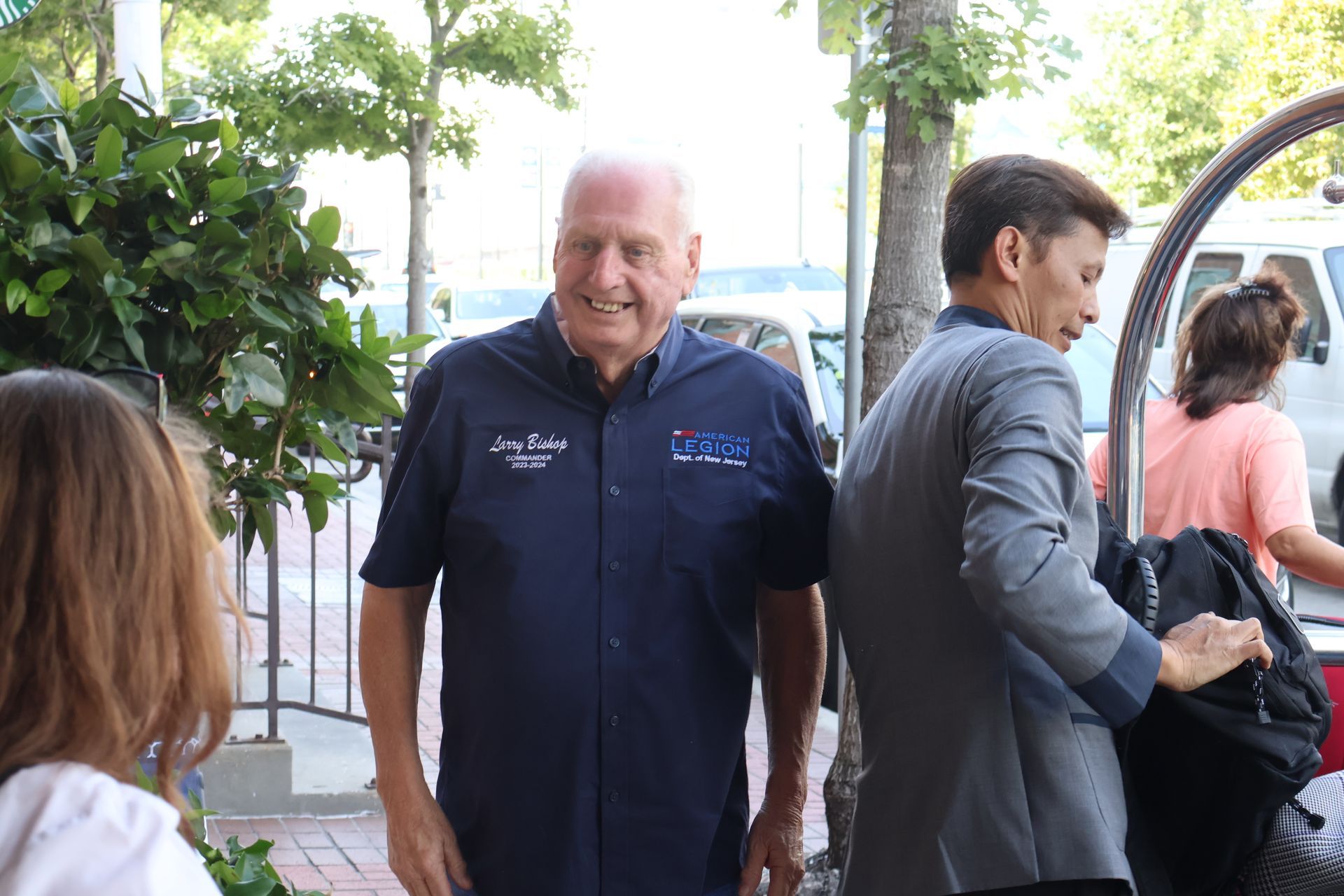 Man in blue shirt smiles outside; another man in a suit looks toward a car.