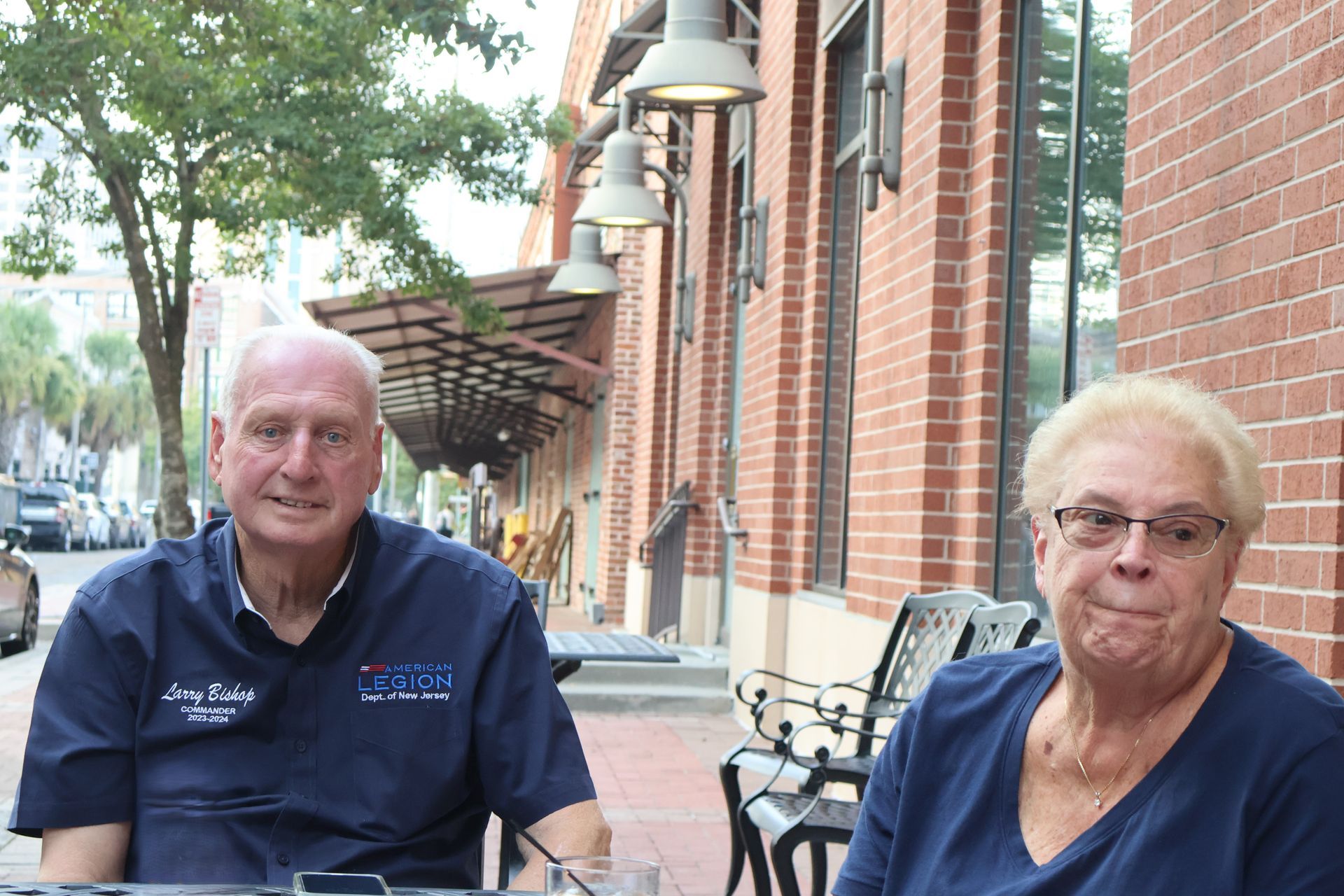 Two people sitting at a table outside a brick building. One wears a blue shirt and the other wears a blue shirt and glasses.