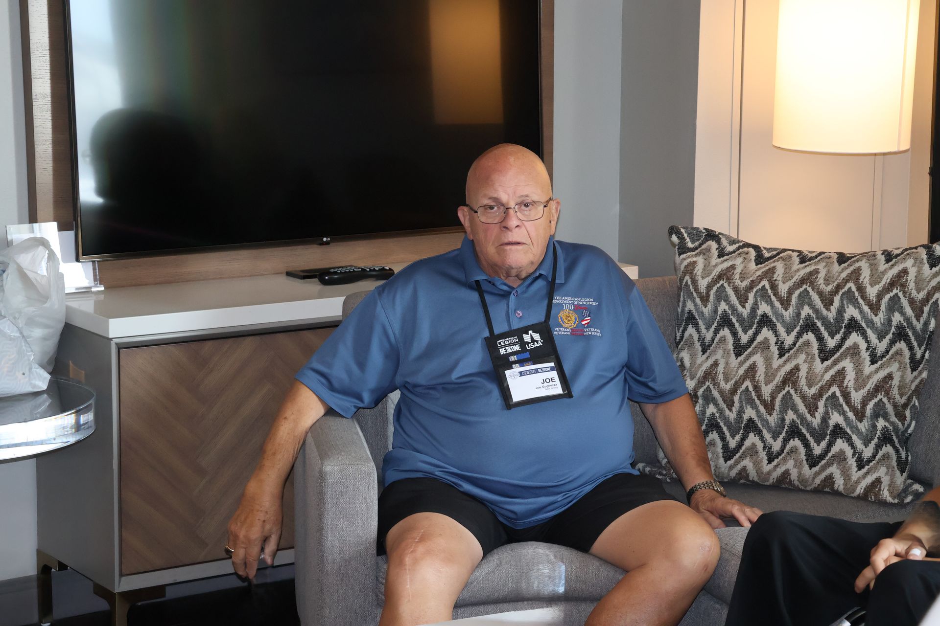 Man sitting in an armchair wearing glasses and a blue shirt, with a name tag, in a room with a TV and lamp.