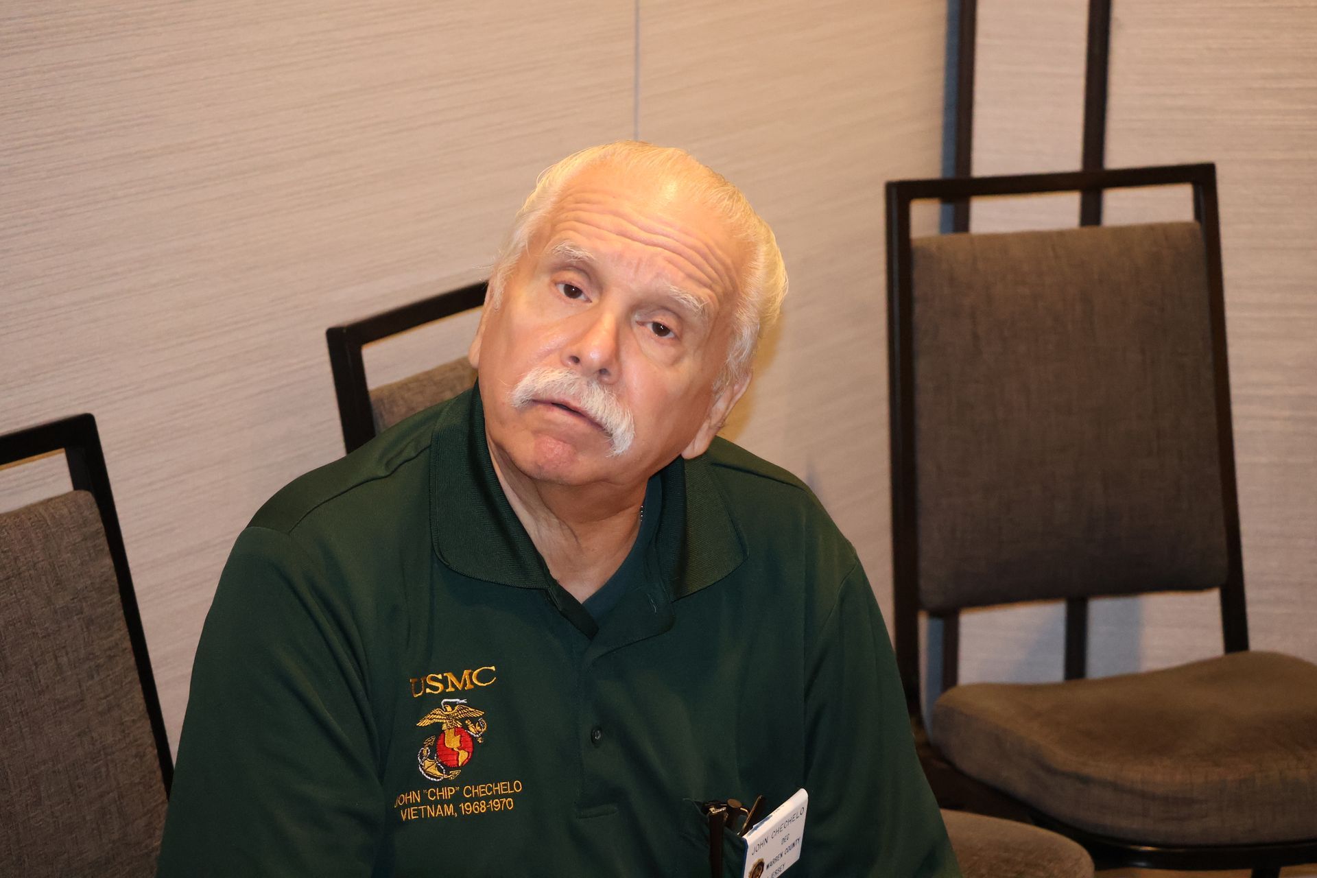 Man in green shirt with mustache, sitting, looking slightly to the side. Behind him are chairs and a wall.