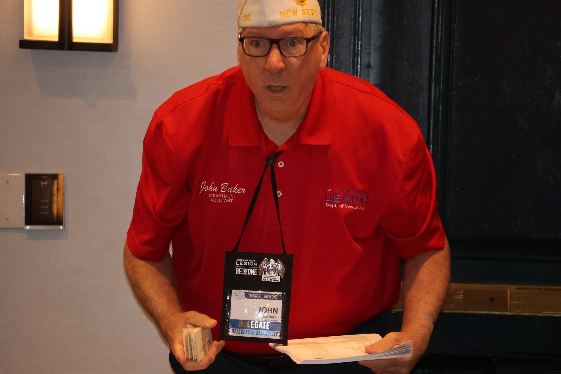 Man in red polo shirt, glasses, and a hat holding a stack of papers and a lanyard with a badge.