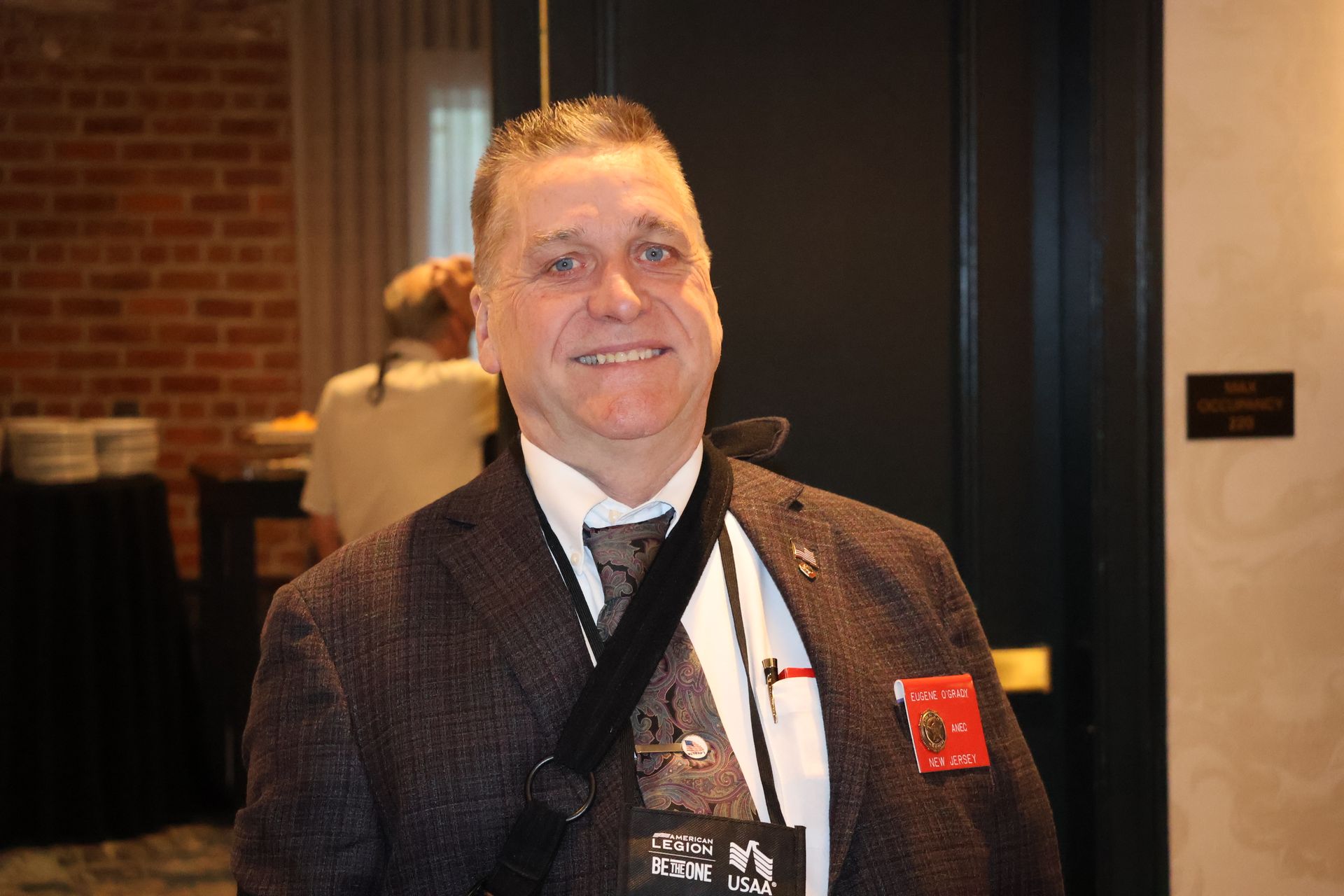 Man in a suit smiles, wearing a lanyard and name tag, standing in a room with a brick wall.