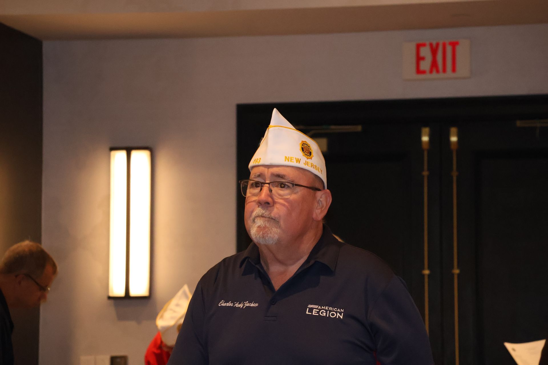 Man in navy shirt and hat, standing indoors near an exit sign.