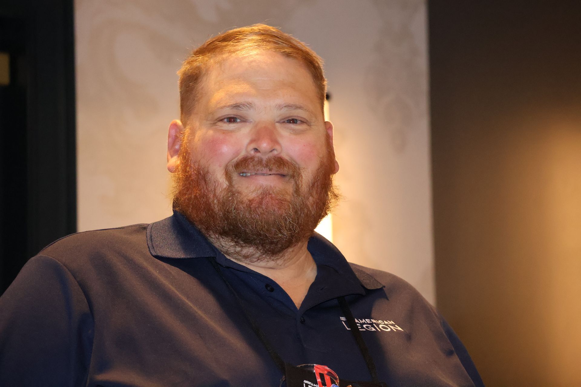 Man with reddish-brown hair and beard smiles, wearing a navy polo shirt and nametag, indoors.
