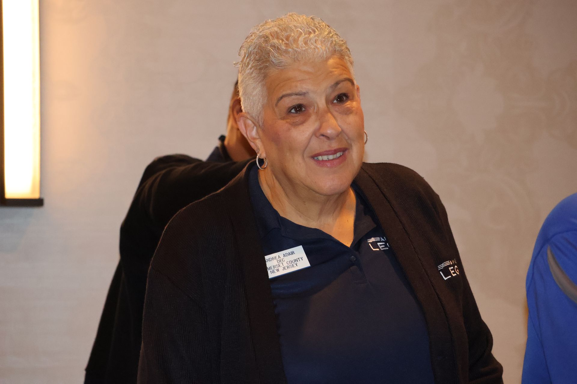 Woman with short white hair smiles, wearing a navy blue shirt and black cardigan in a conference setting.