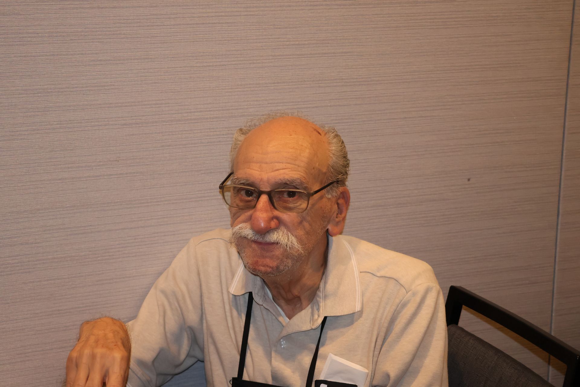 Man with glasses and mustache smiles, wearing a light-colored shirt, seated. Gray wall background.