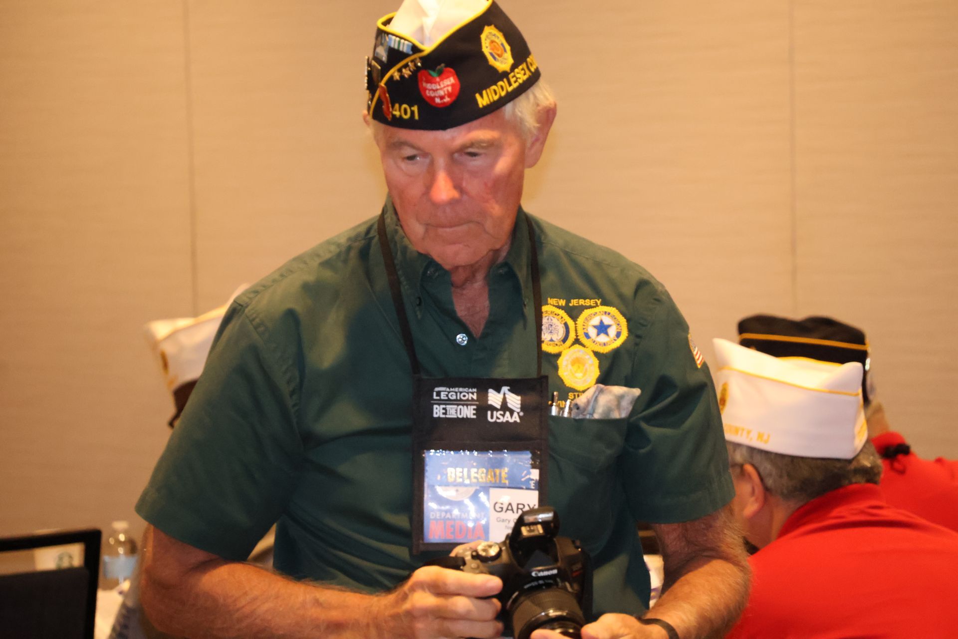 Man in a green shirt and veteran's cap holding a camera. He is in a meeting room with others.