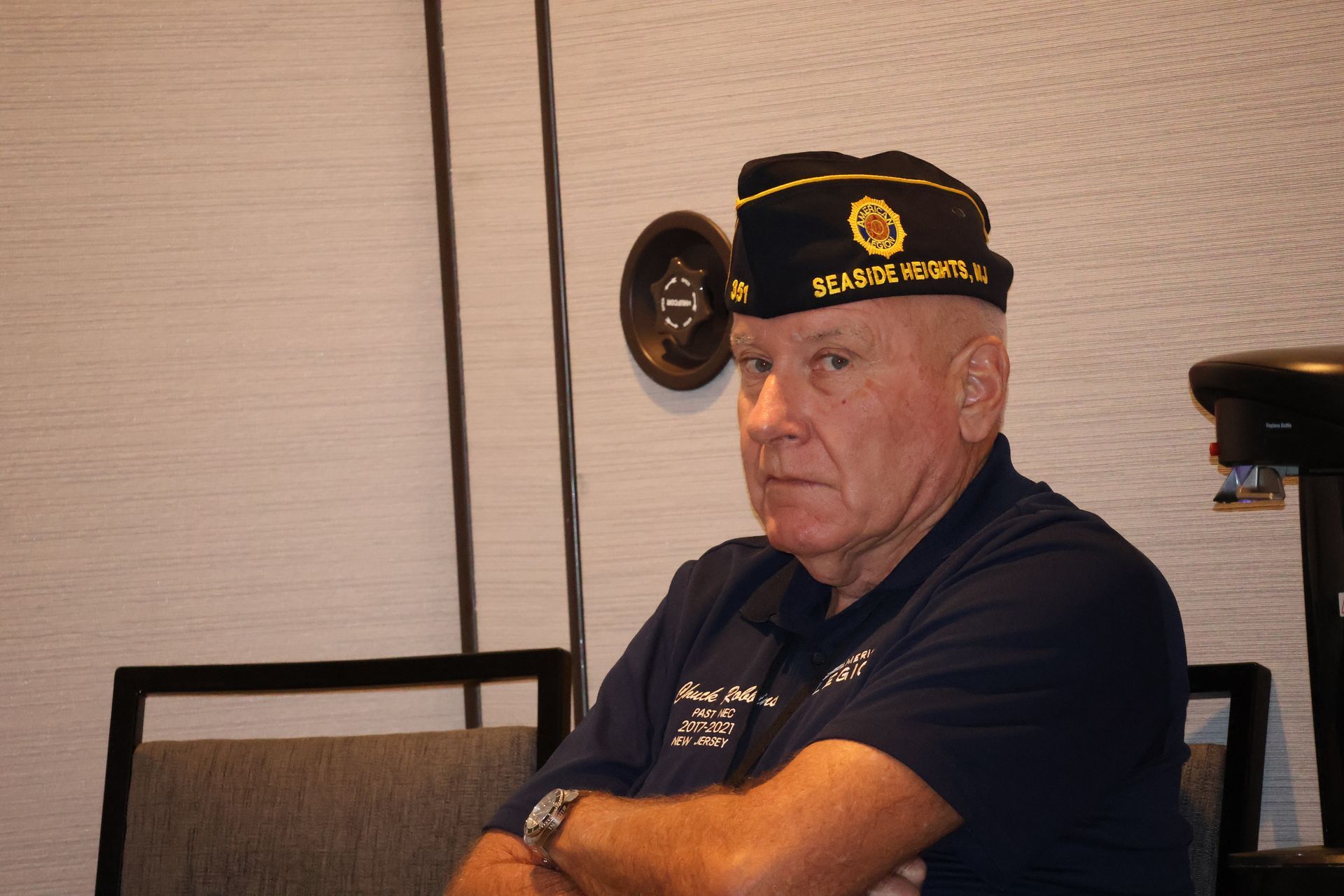Man wearing a veteran's hat and blue shirt, seated with arms crossed, looking at the camera.