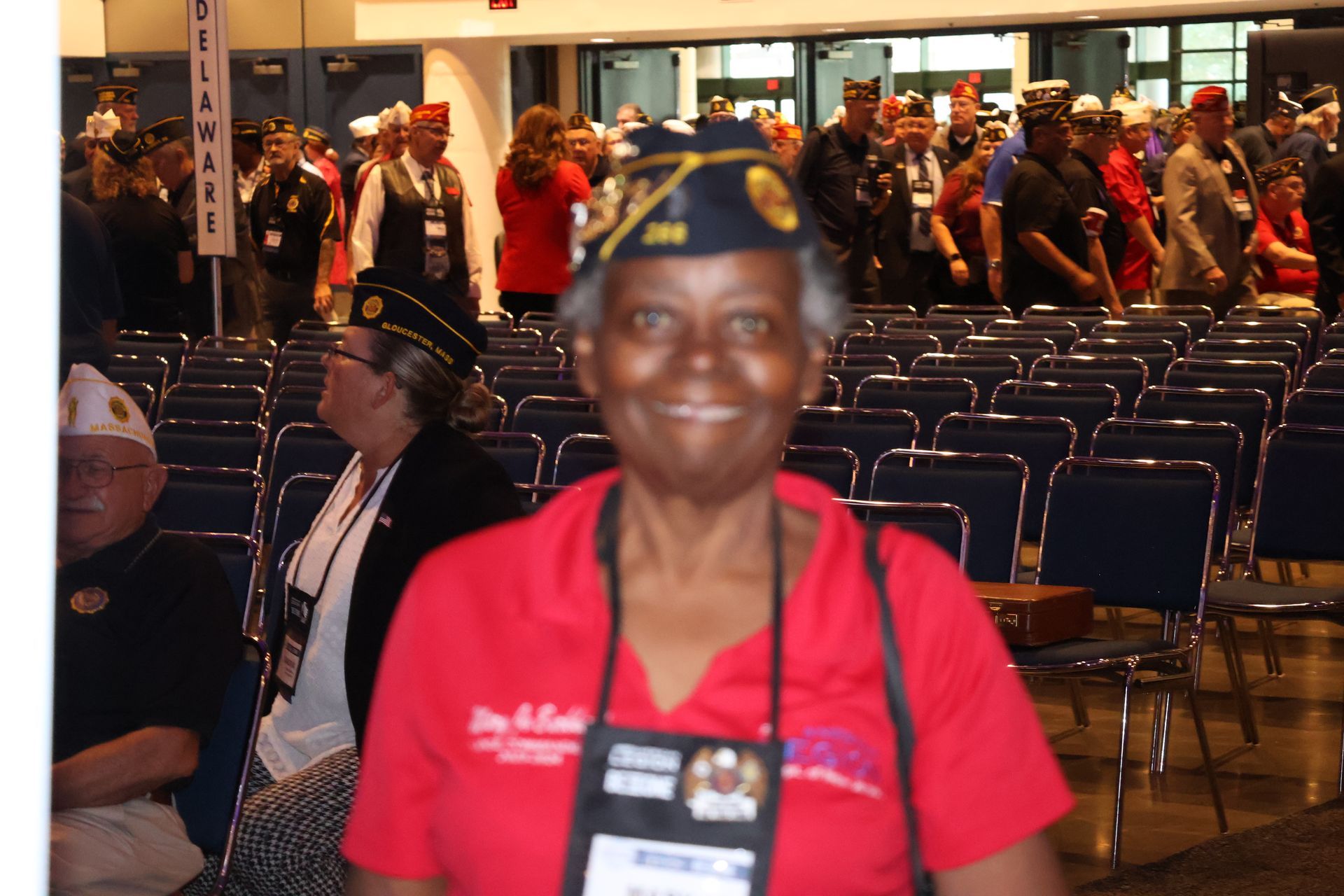 Woman in red shirt and hat smiles at a veterans' conference with others in background.