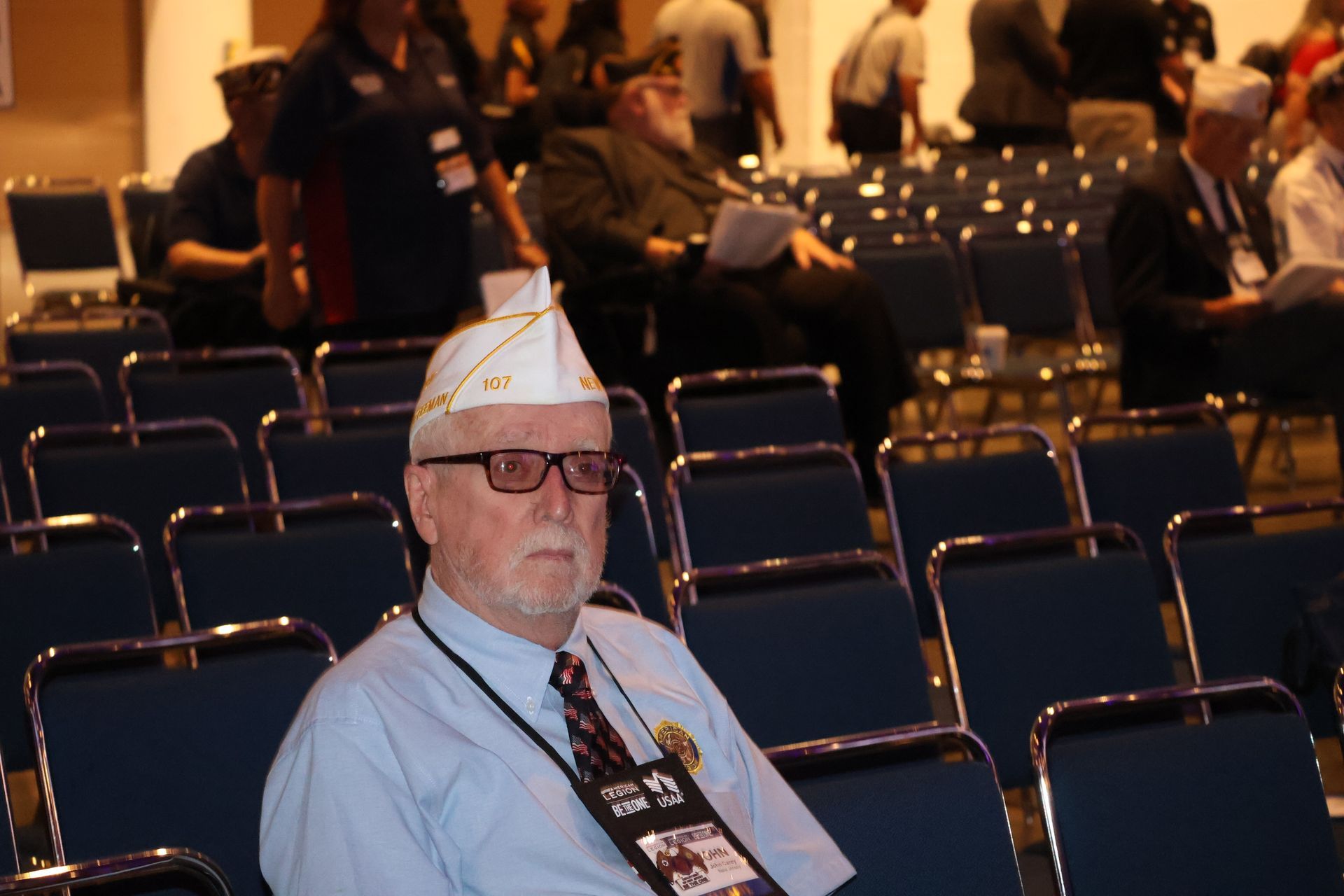 Man in veteran's cap seated in an auditorium, wearing a lanyard. Others sit behind him.