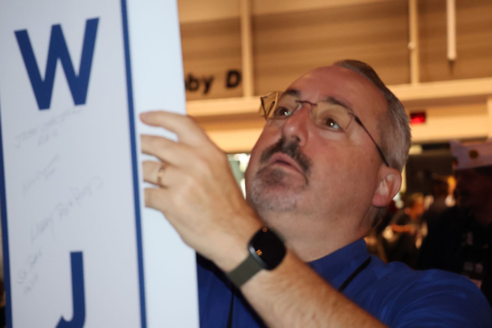 Man in glasses and blue shirt looking up at a white sign with blue lettering; he is touching the sign.