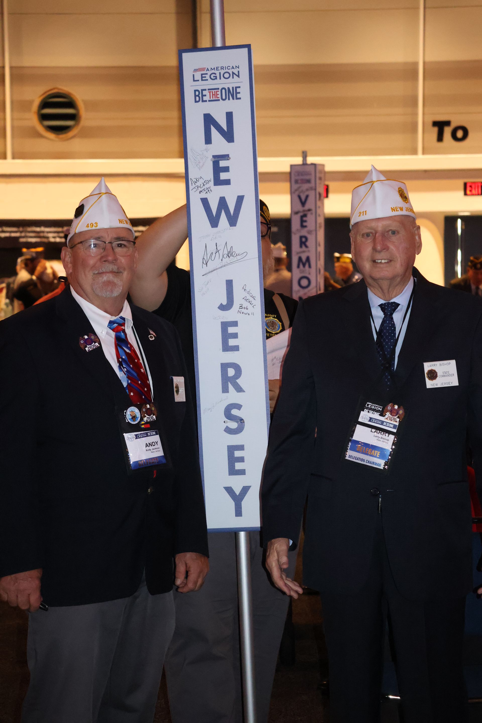 Two men in navy blazers and hats stand next to a sign that says 