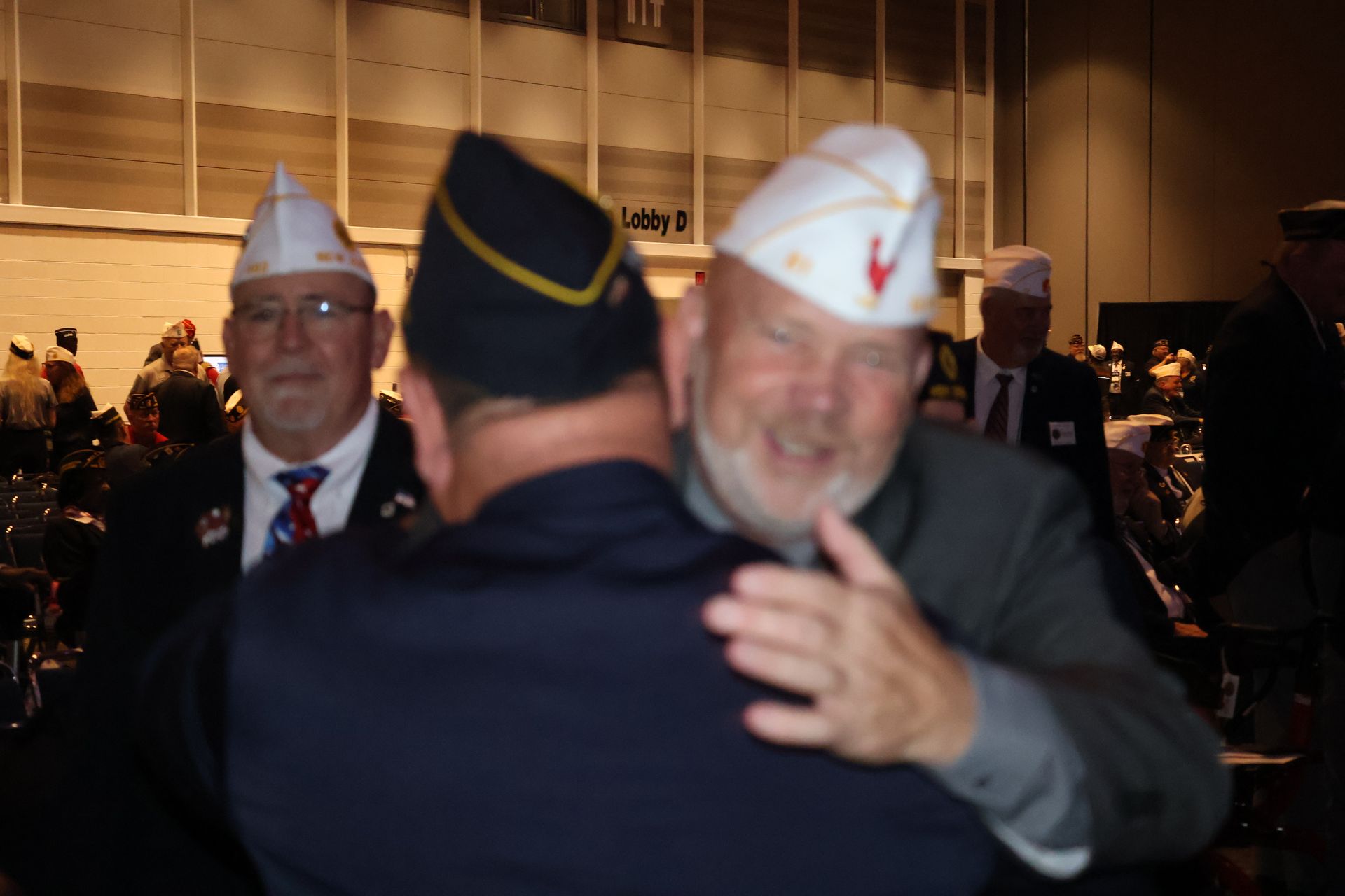 Three men in veteran hats, one hugging another. Two smile. Interior setting.