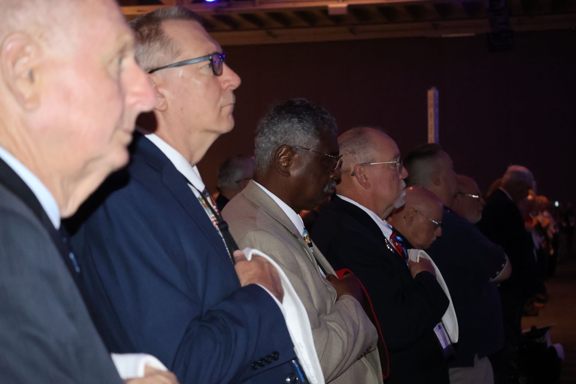 Men in suits standing and looking forward in a dimly lit room, possibly at an event.