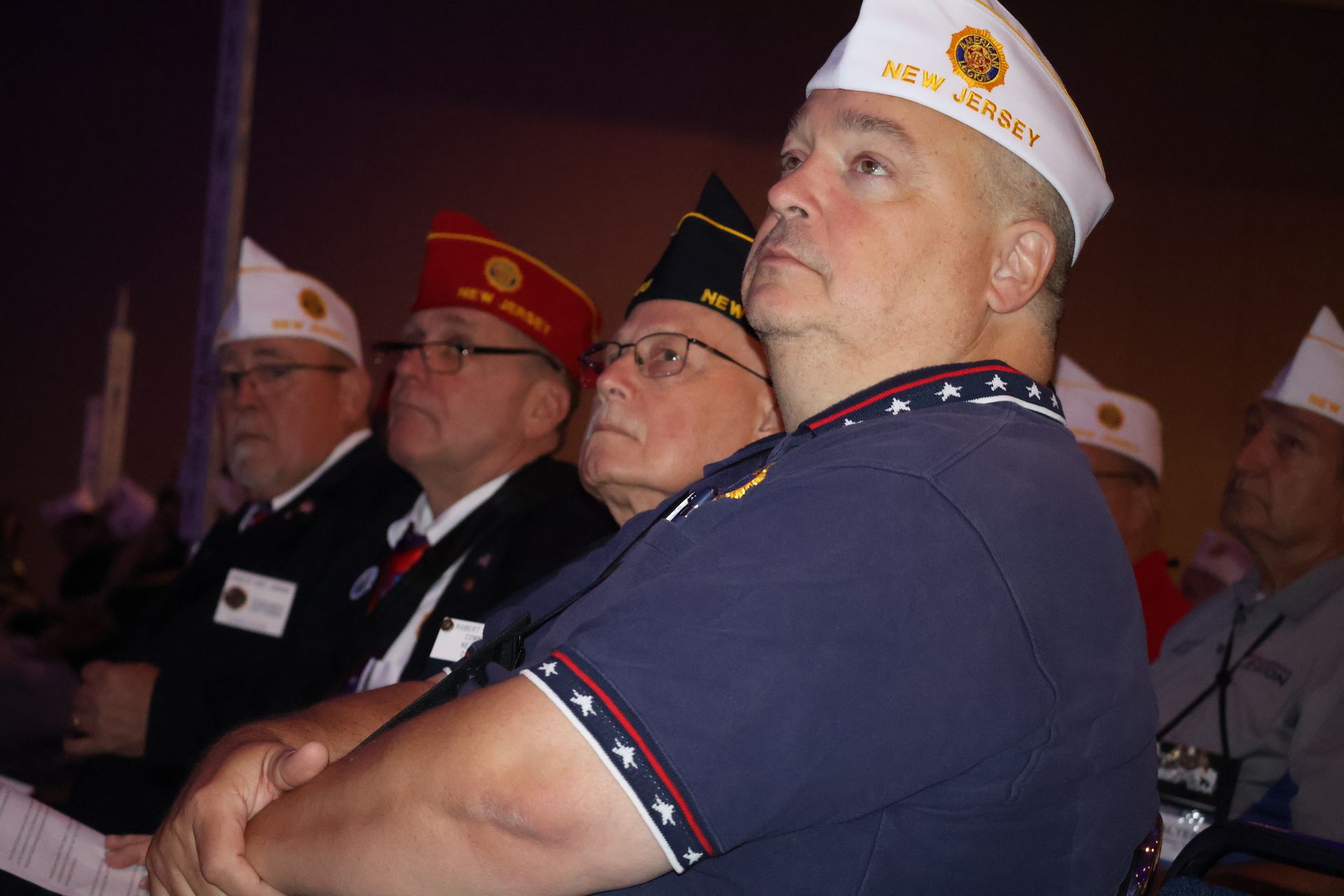Men in veteran hats listen attentively, arms crossed, in a meeting setting.