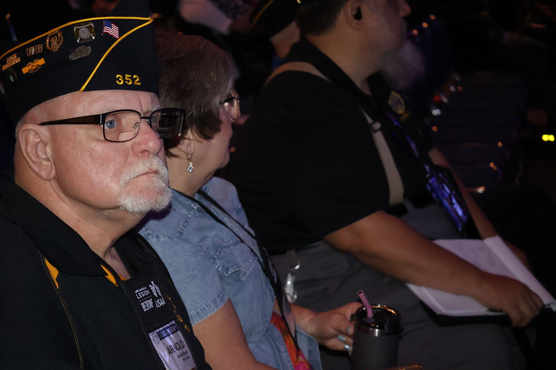 Man in a veteran's cap and jacket, looking to the side, seated next to a woman and another person.