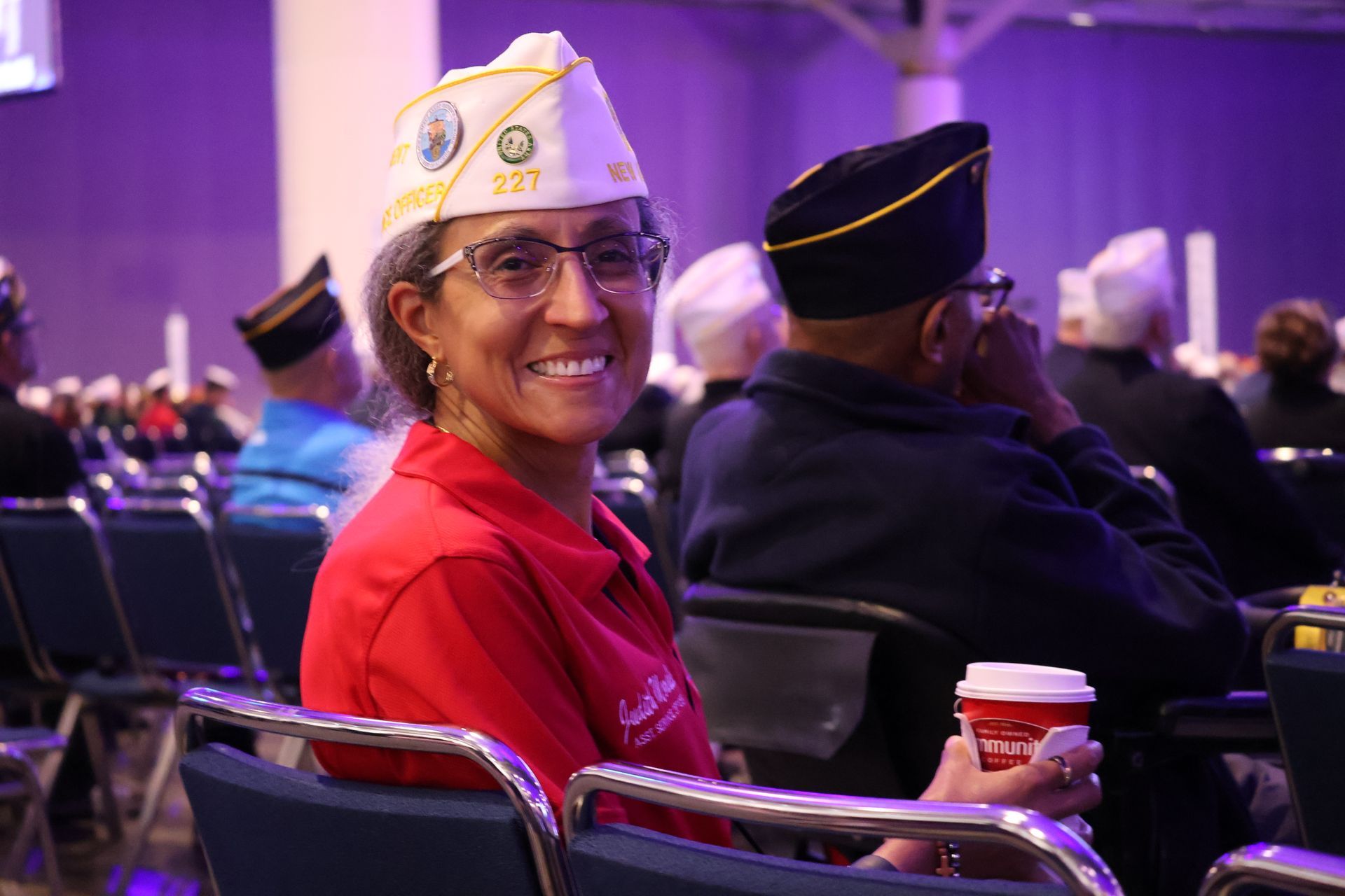 Woman in red shirt and veteran cap smiles, holding a coffee cup, seated at an event.