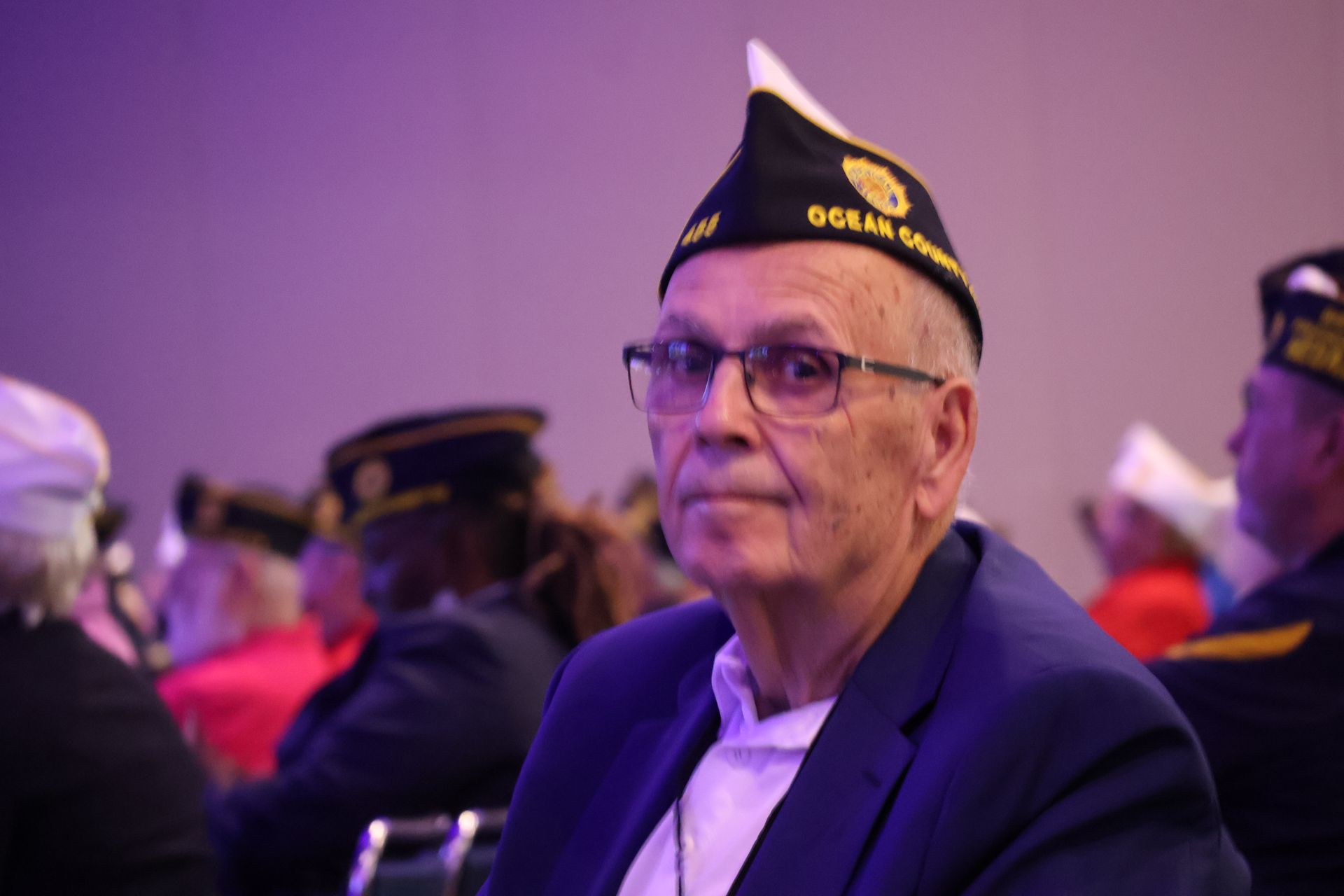 Man wearing a veteran hat and glasses, seated, looking forward, surrounded by others in a room.