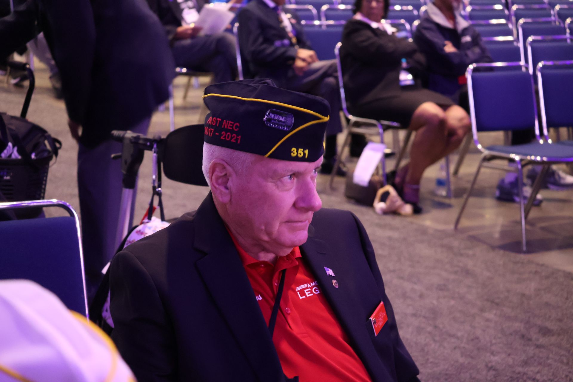 Man in veteran's cap, red shirt, and black blazer, seated in a room, looking right. People in background.