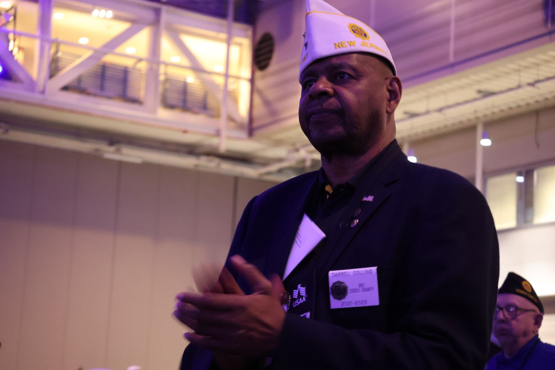 Man in navy blazer and veteran's cap clapping in a brightly lit indoor space.