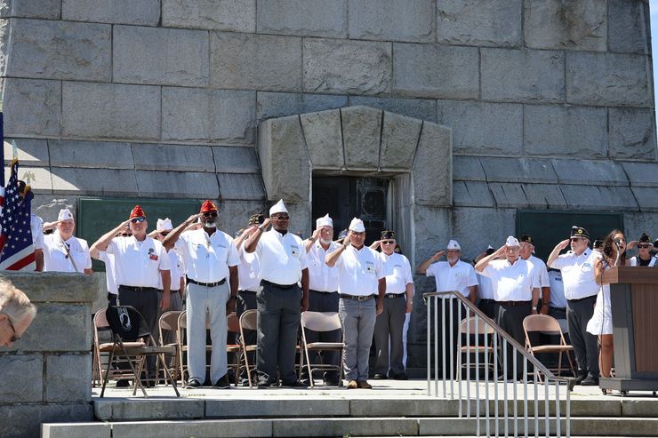 Veterans saluting at a memorial service, standing on a stone platform, sunny day.