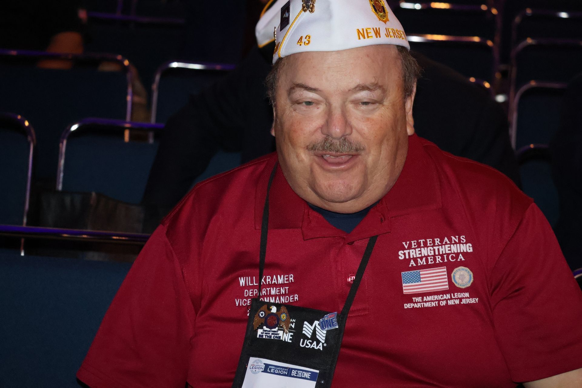 Man wearing a veteran's hat and red shirt. Smiling, sitting indoors with an ID badge.