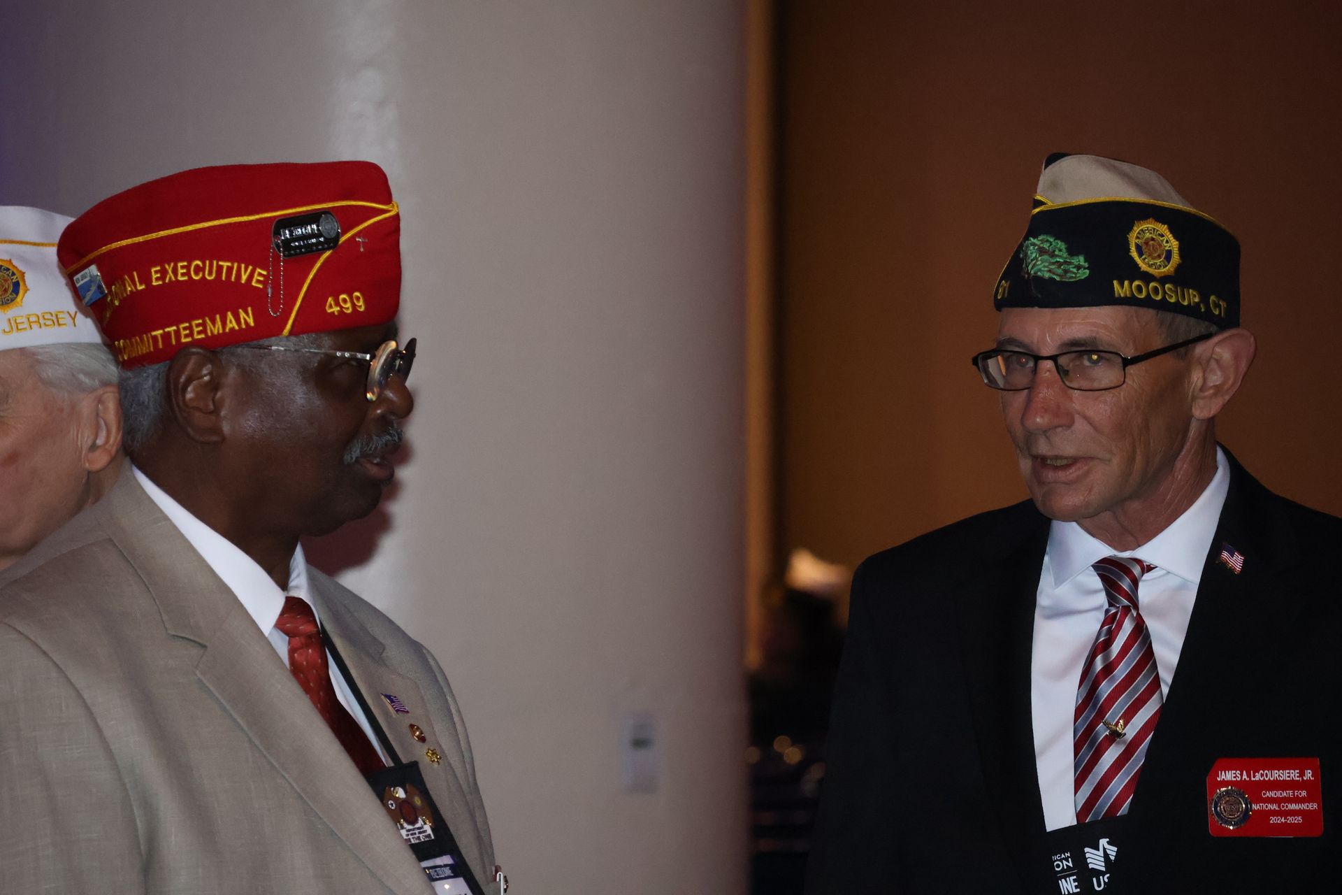 Two men in suits and veteran hats converse indoors.