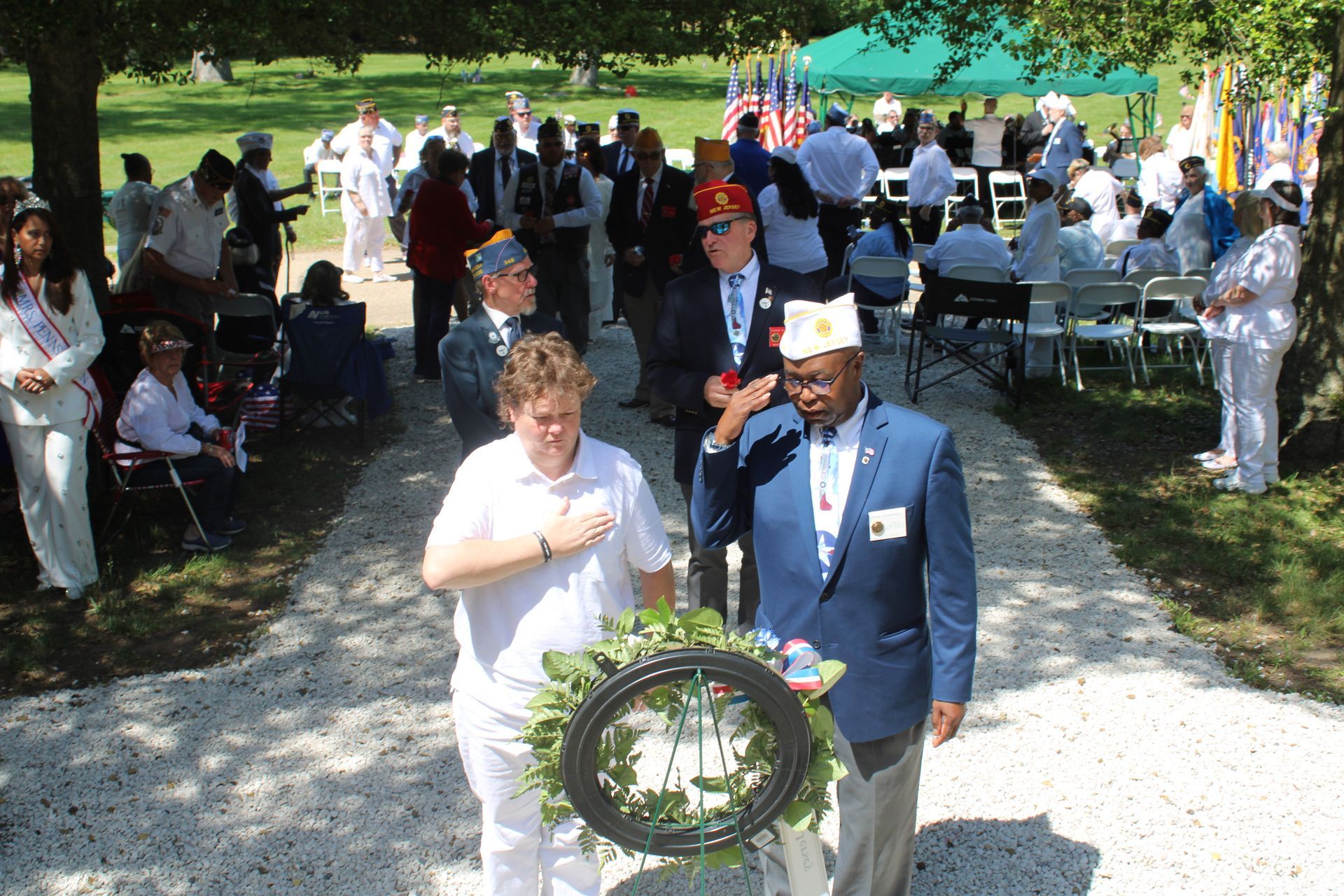 People at an outdoor memorial ceremony gather around a wreath in a park.