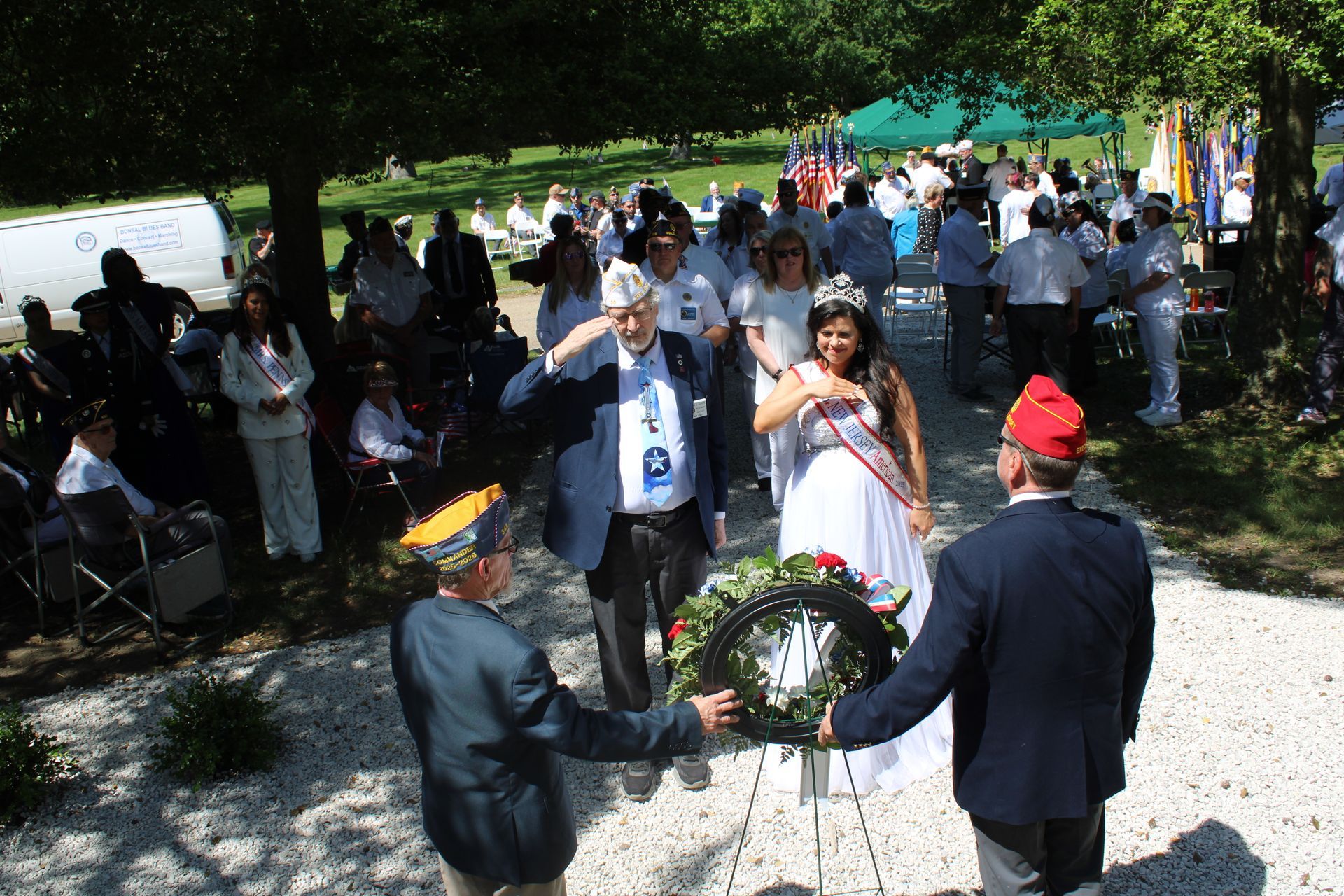 Mourning ceremony outdoors, with people gathered around a wreath and a man in a red cap standing nearby