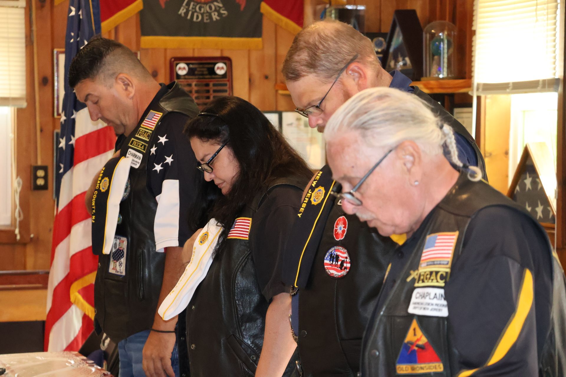 Group of people, wearing vests, bowing heads. Indoors with flags and wooden walls.