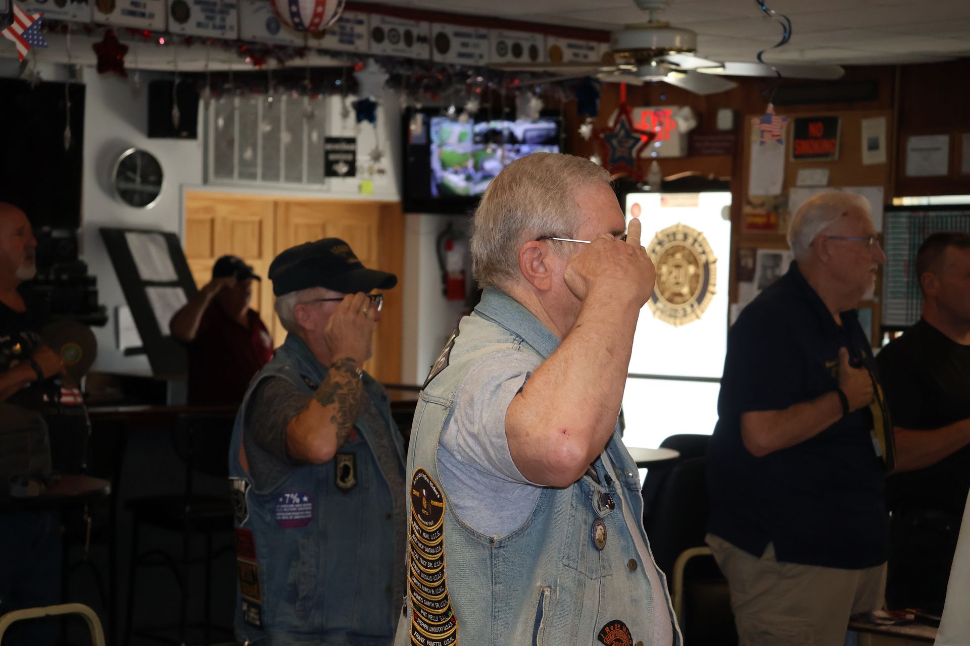 Men in a club, saluting; some wearing vests with patches. Inside with bar and patriotic decorations.
