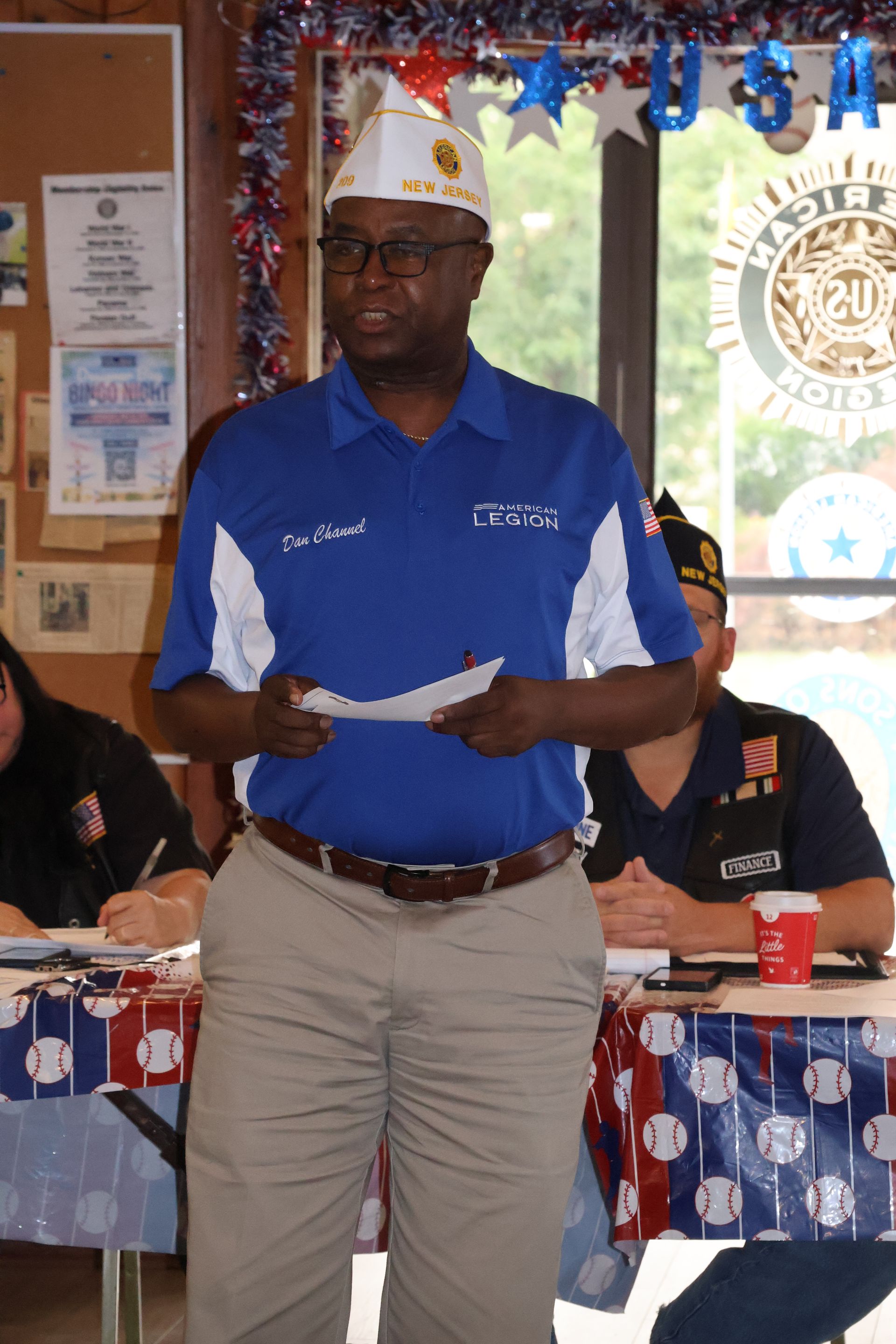 Man in blue shirt and khaki pants speaks at a patriotic event, wearing a service cap, holding papers.