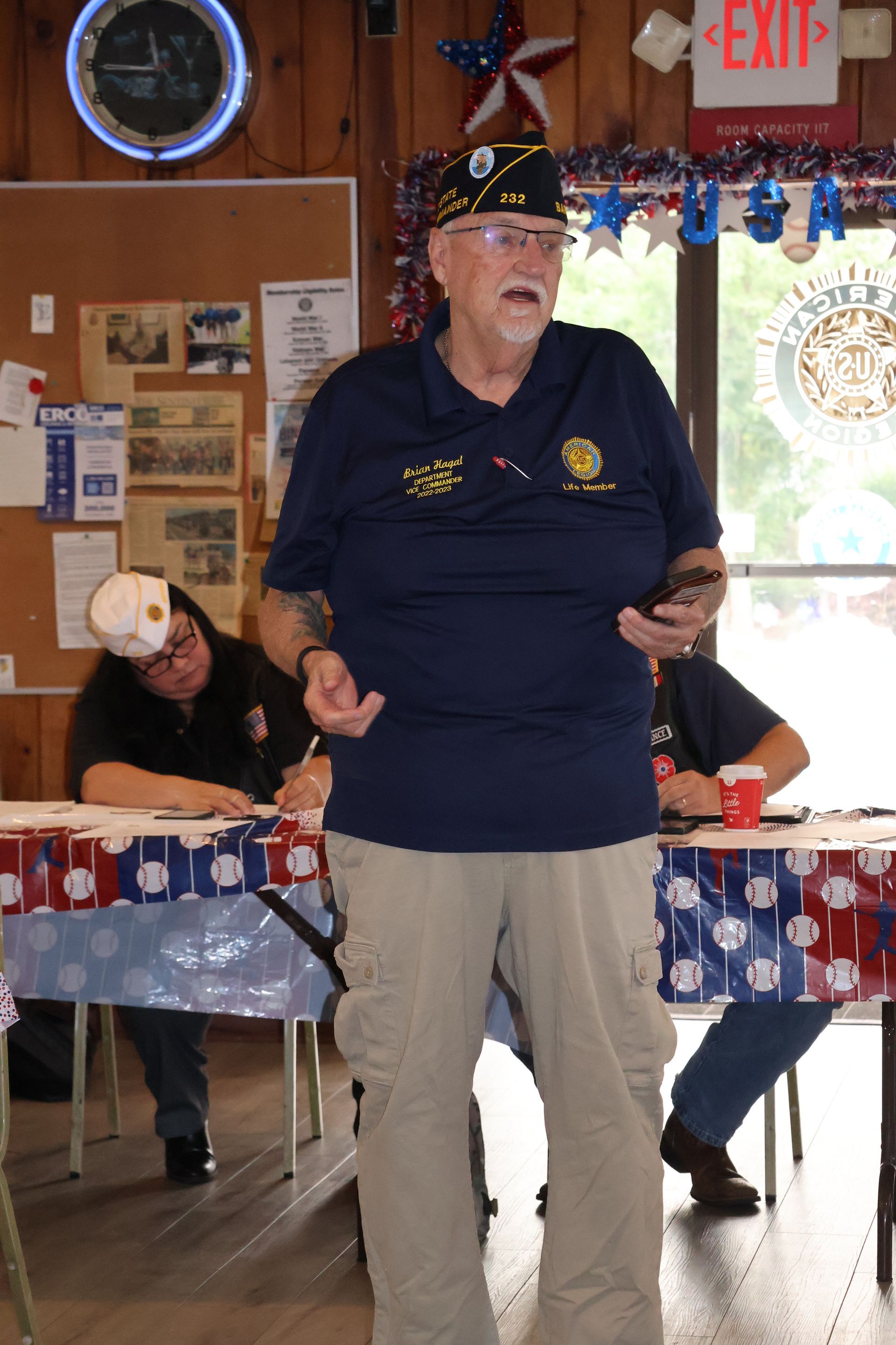 Man in a blue shirt and khaki pants speaks at a patriotic event. Tables with red, white, and blue decor are in view.