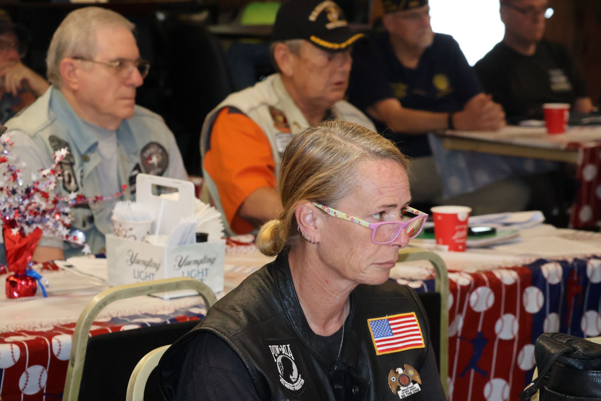 People seated at a table, some wearing veteran attire. A woman with blonde hair and pink glasses is in the foreground.