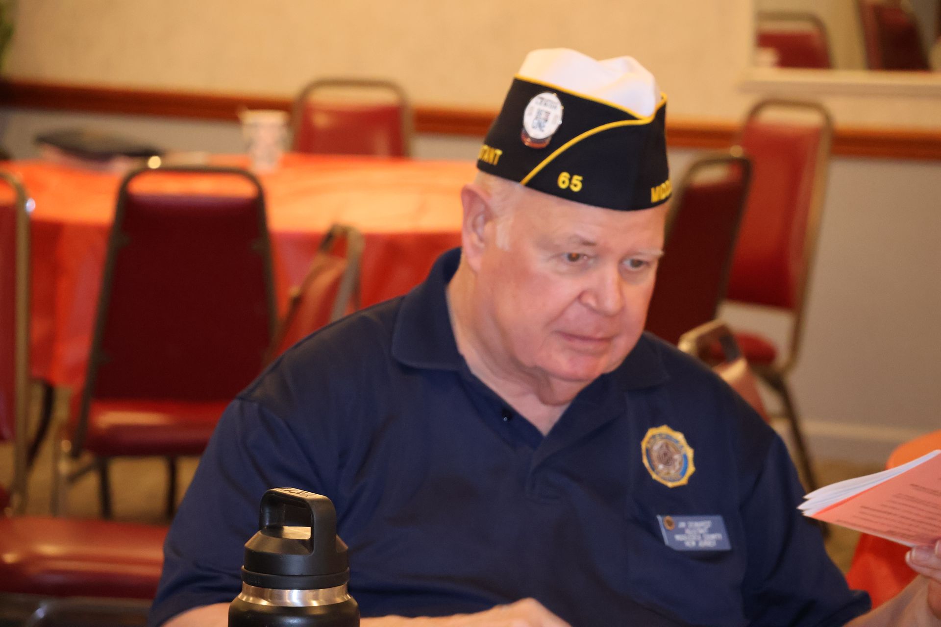 Man in navy blue shirt and cap with insignia, seated at table, looking at papers.