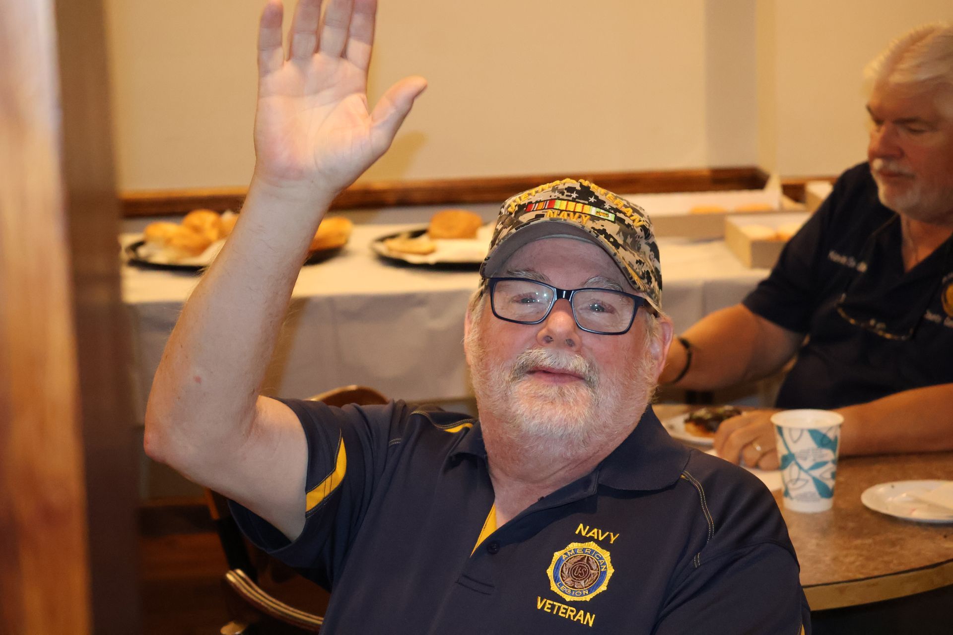 Man in navy shirt and camo cap raises hand, smiles at a gathering.