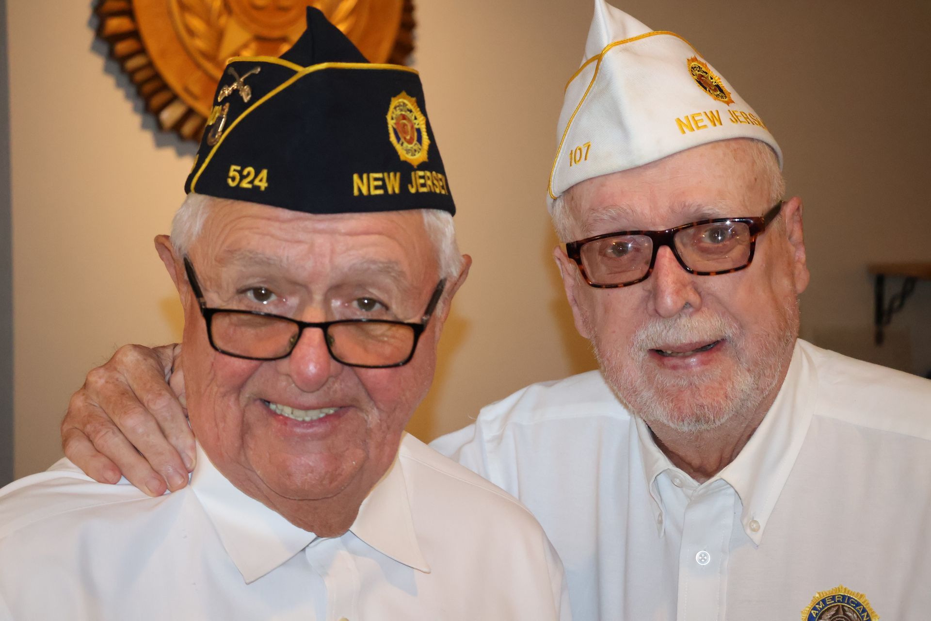 Two men in veteran hats, smiling, indoors. One has a dark blue cap, one has a white cap.