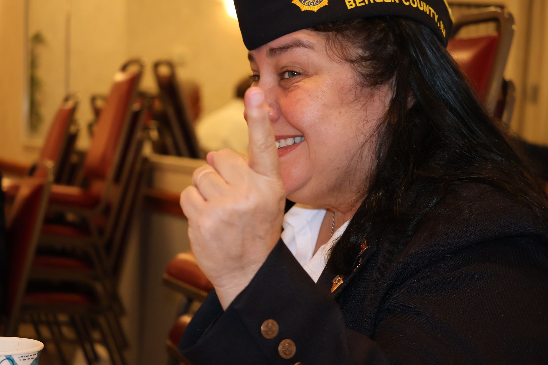 Woman in a navy blazer and hat smiles, pointing upward with her index finger.