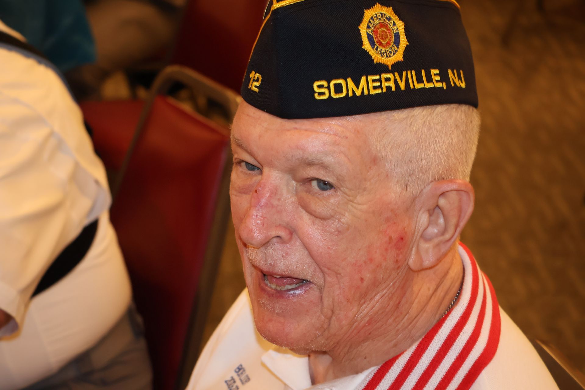 Man wearing a Somerville, NJ cap, and red, white, and blue sash speaks; indoor setting.