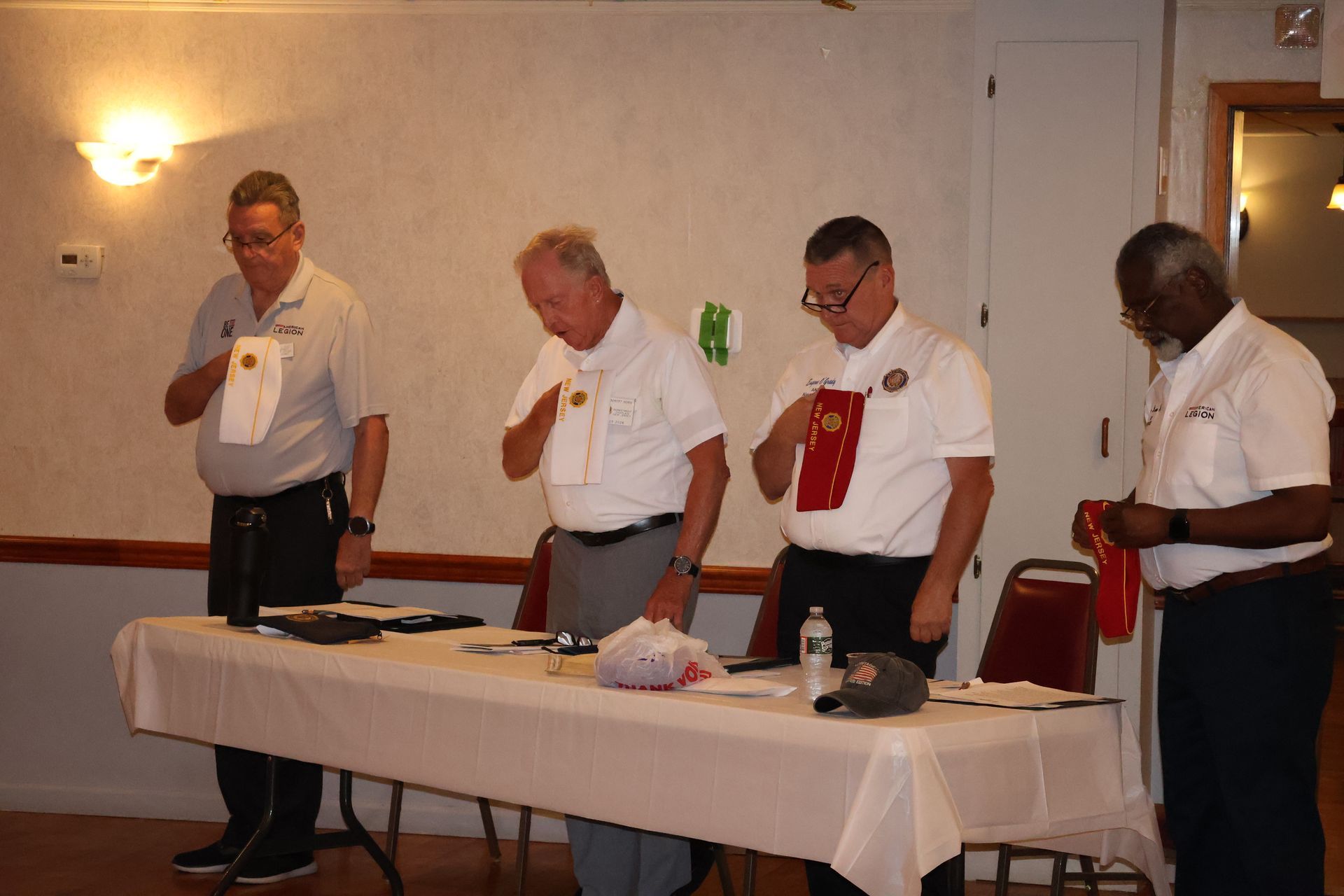 Four men at a table, holding papers, with hands over their hearts. Inside a room with dim lighting.