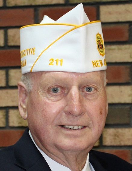 Man wearing a white and gold military-style hat, navy suit. He smiles in front of a brick wall.