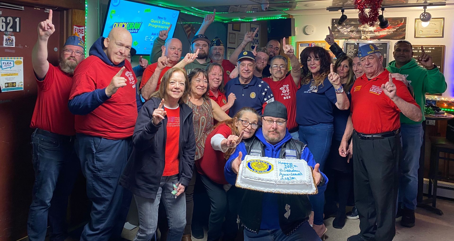 Group of people celebrating, holding a cake. They are in a bar setting, some raising their hands.
